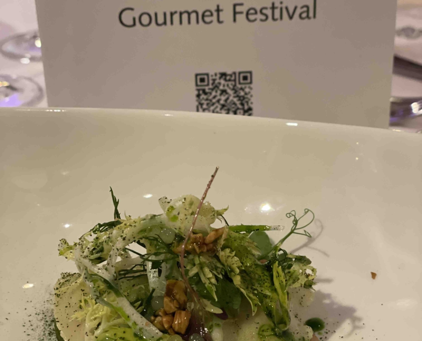 Ein grüner Salat mit Öl, Krabben und Nüssen liegt in einer weißen Schale. Dahinter ist die Menükarte des Abends zu sehen.